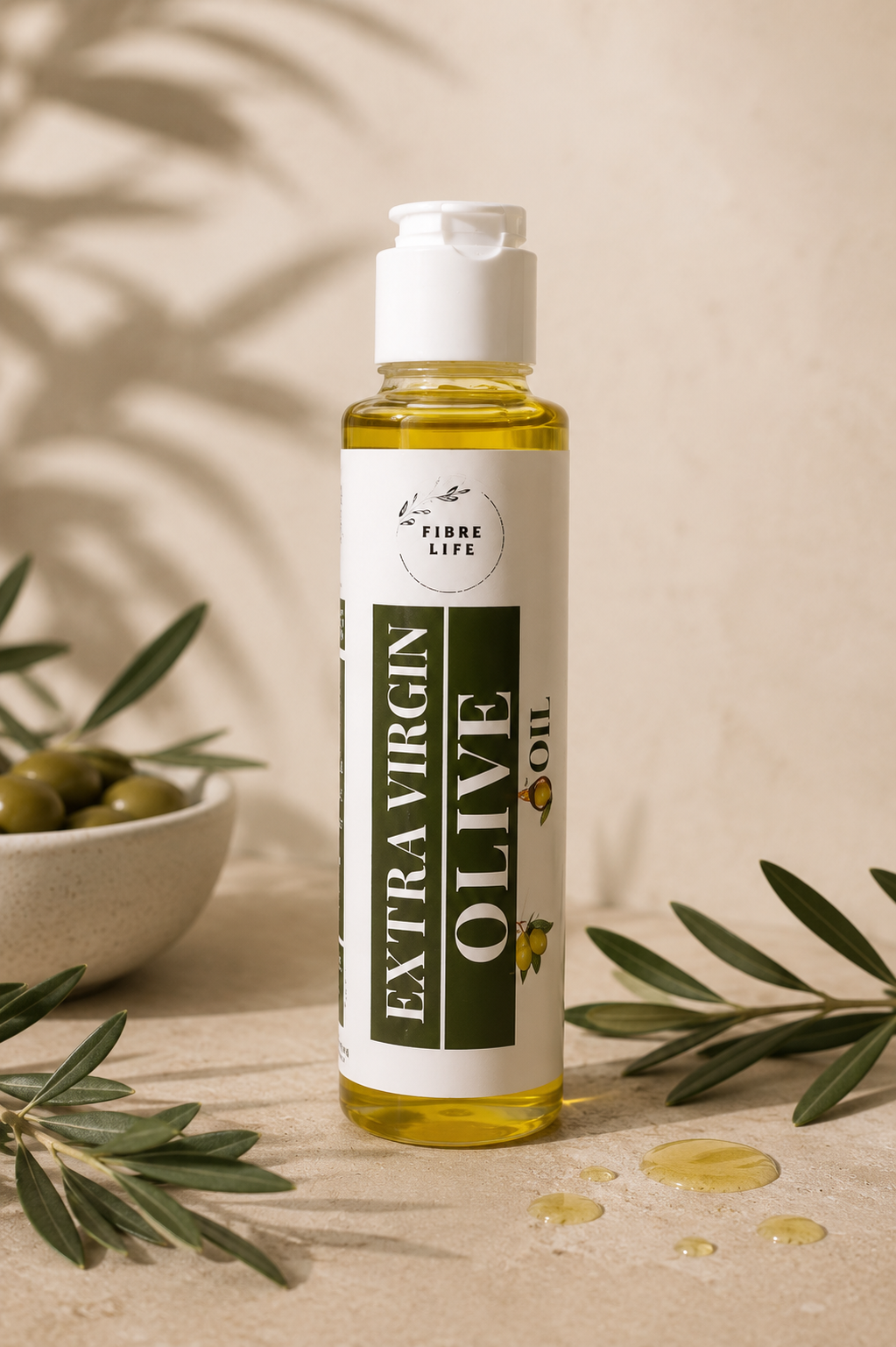 A bottle of Fibre Life Extra Virgin Olive Oil stands on a beige surface with olive branches and a bowl of olives around it. The bottle is clear, showing the golden oil inside, with a white cap and a white label featuring dark green text.